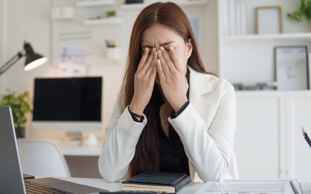 A professional overwhelmed at her desk with her hands over her face